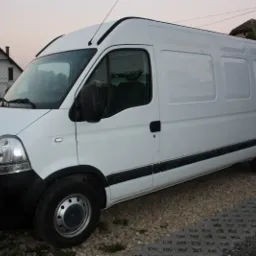 Biały samochód dostawczy Renault Master sfotografowany z boku na tle domu z dwuspadowym dachem i szarym niebem.