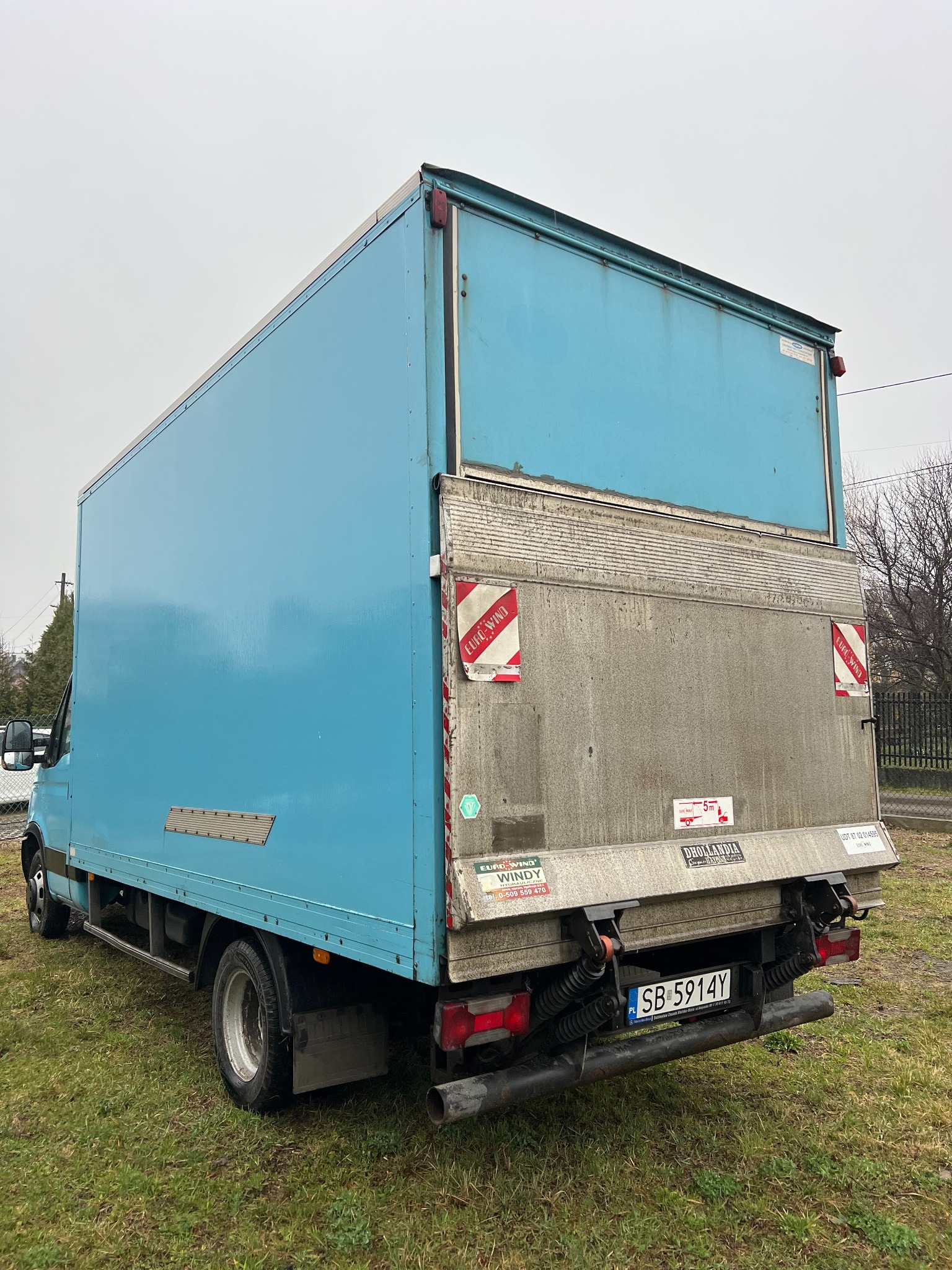 Niebieski samochód dostawczy z otwartą klapą załadunkową, widok z tyłu na trawiastym podłożu. Widoczne naklejki i tablica rejestracyjna SB 5914Y.