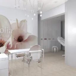 Jasne wnętrze gabinetu kosmetycznego z białymi meblami, przezroczystymi krzesłami, lampą na statywie i fototapetą z motywem magnolii.