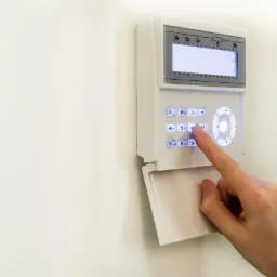 Biały panel alarmowy na ścianie z podświetlanymi przyciskami, obsługiwany przez osobę wciskającą przycisk '6'. Na wyświetlaczu widoczna data i godzina.