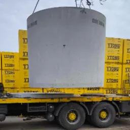 Betonowy krąg studzienny z napisem 'BREJNAK' transportowany na platformie samochodu ciężarowego, obok palet z bloczkami YTONG.
