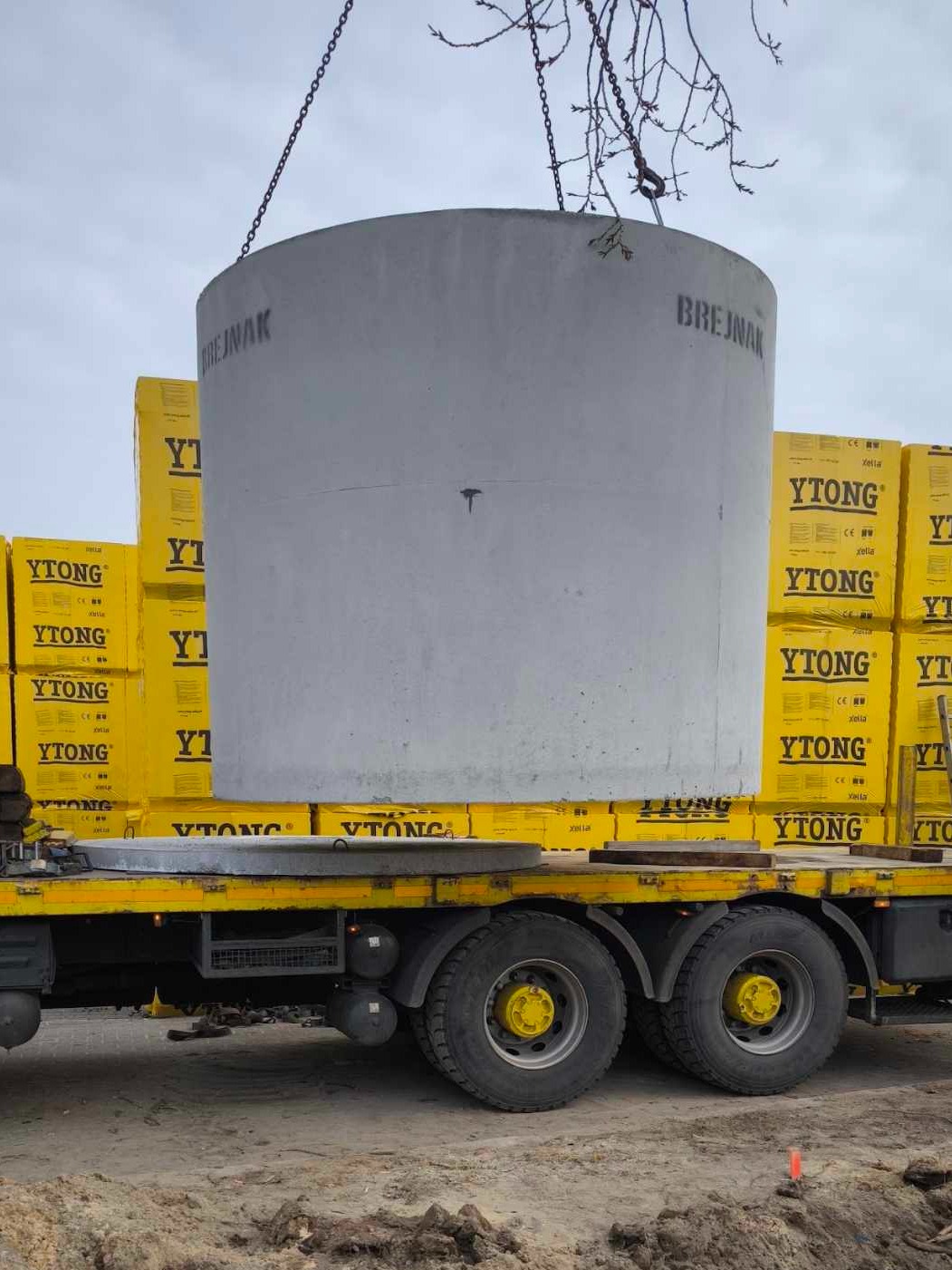 Betonowy krąg studzienny z napisem 'BREJNAK' transportowany na platformie samochodu ciężarowego, obok palet z bloczkami YTONG.