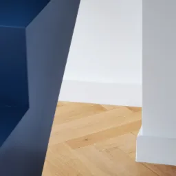 Parkiet w jodełkę, pomalowana na biało ściana i fragment zabudowy w kolorze granatowym, minimalistyczne wnętrze.