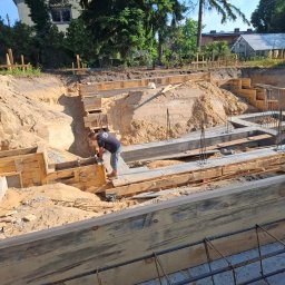 Property Investments Maciej Morawski - Budowa fundamentów domu: szalunki z desek, zbrojenie stalowe, wykop ziemny. Pracownik w tle. Widok z góry na plac budowy w słoneczny dzień.