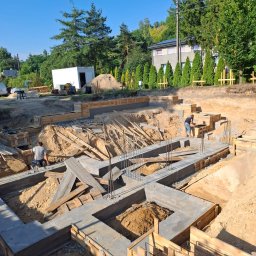 Property Investments Maciej Morawski - Fundamenty domu w trakcie budowy: betonowe ściany szalunkowe, zbrojenia stalowe, drewniane elementy konstrukcyjne. Dwóch pracowników w trakcie prac. Widok z góry, słoneczny dzień.