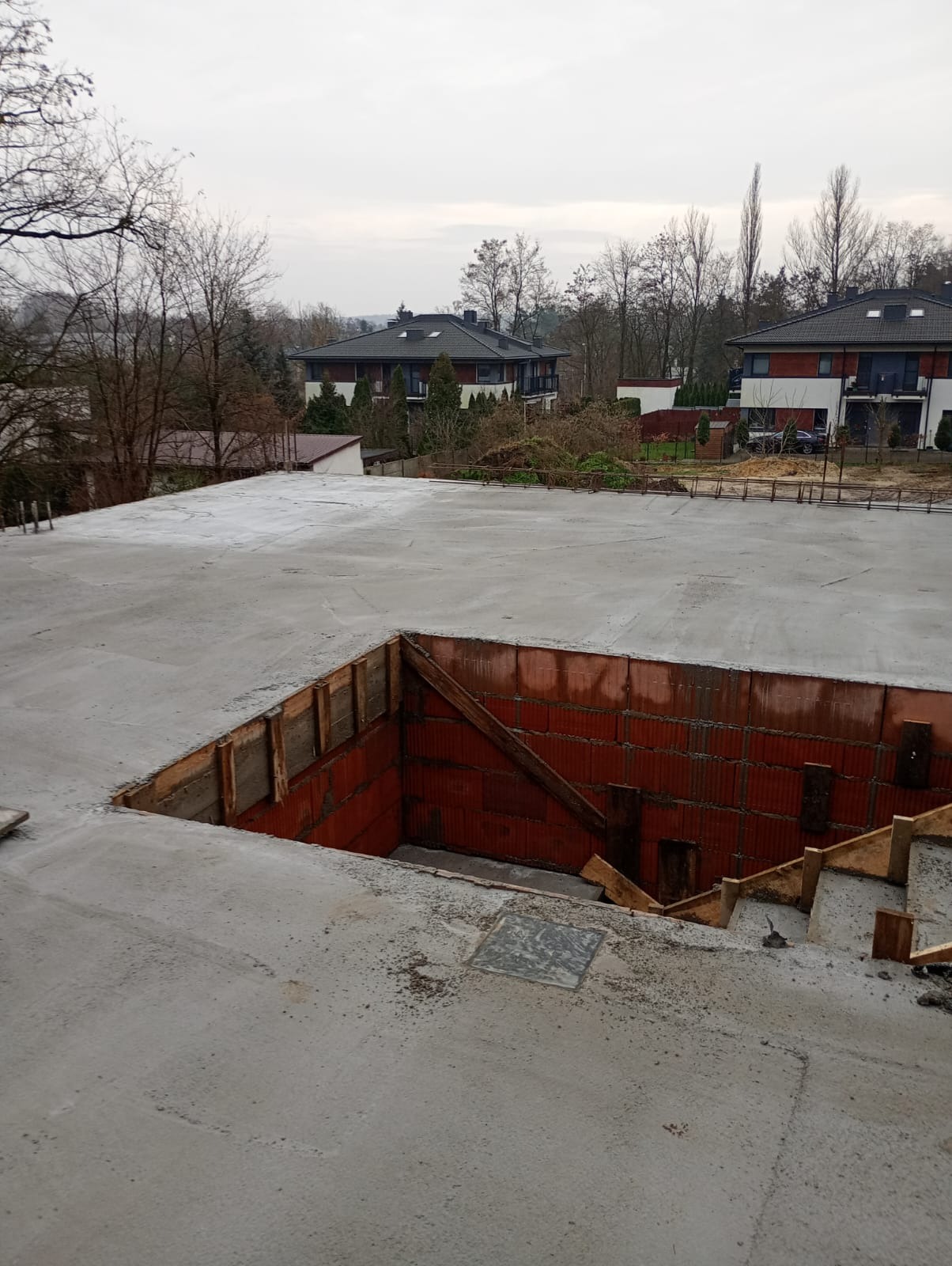 Surowy betonowy strop z otworem na schody, widoczne ceglane ściany i drewniane szalunki. W tle domy jednorodzinne i drzewa w jesiennej scenerii.