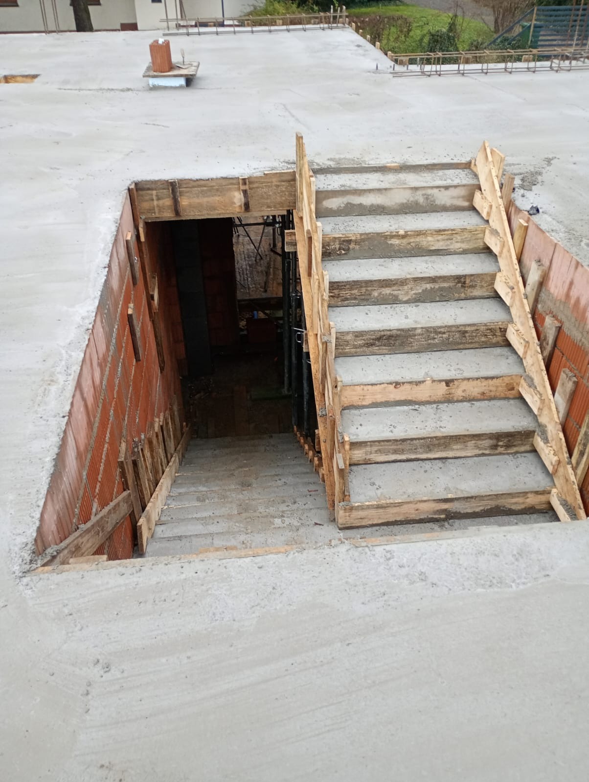 Surowe betonowe schody z drewnianym szalunkiem prowadzące w dół, widoczne ceglane ściany i betonowa płyta stropowa. Etap budowy w surowym stanie.