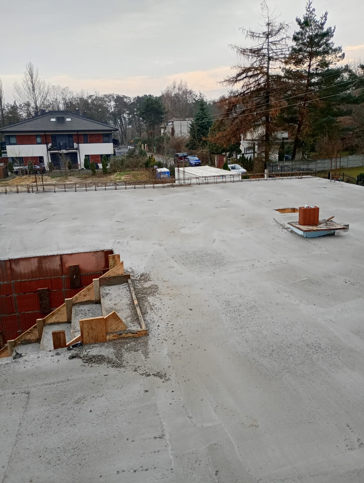 Wylany betonowy strop domu jednorodzinnego w trakcie budowy, widoczne szalunki schodów i komina. W tle zabudowa jednorodzinna i drzewa.