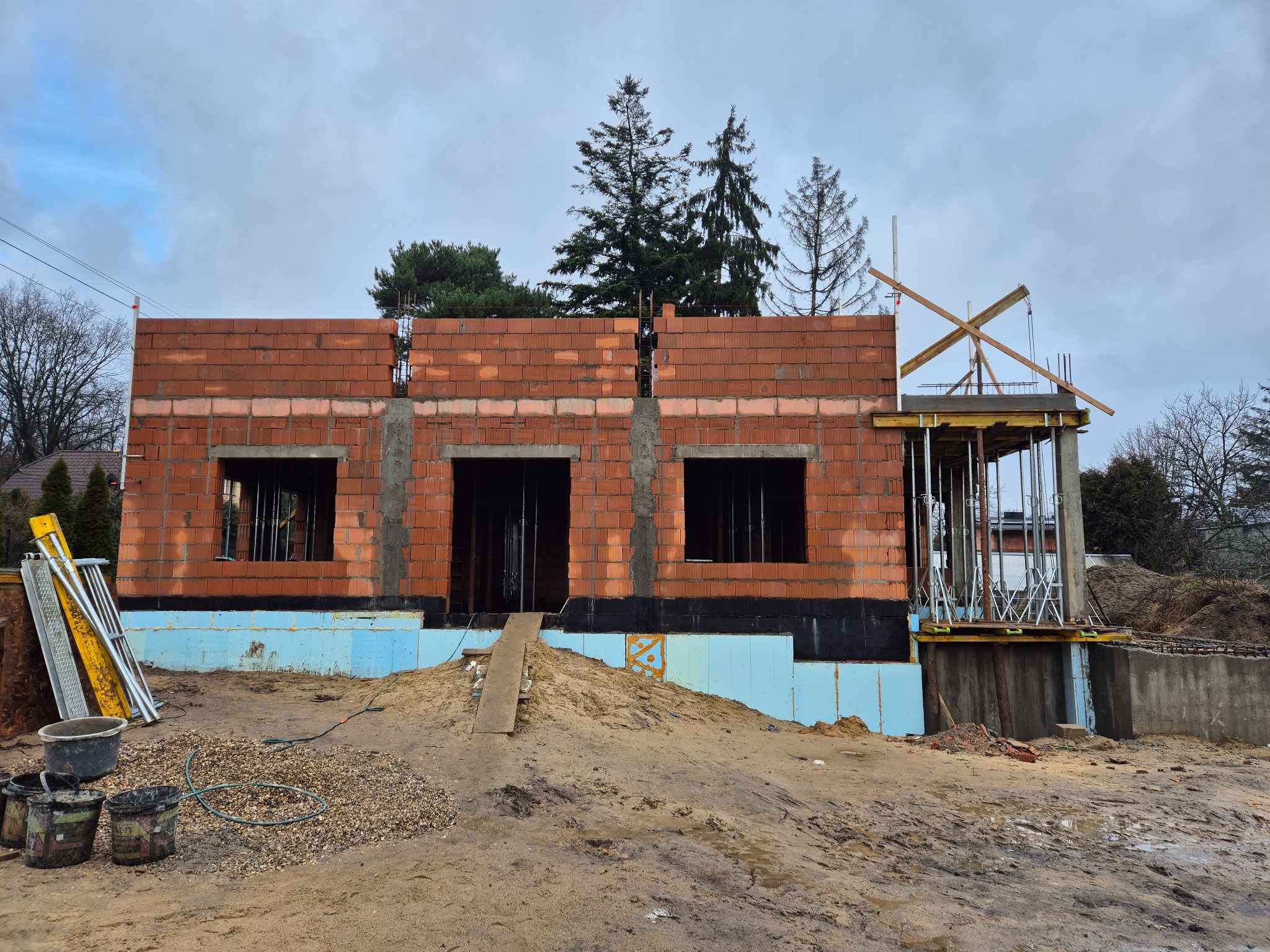 Wznoszenie domu z czerwonej cegły: ściany parteru z otworami okiennymi, ocieplenie fundamentów, szalunki na betonowym fundamencie. Widoczne narzędzia i materiały budowlane.
