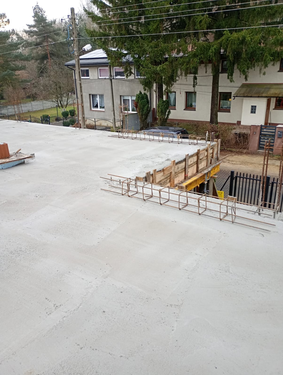 Zalany betonowy strop z widocznym zbrojeniem i szalunkiem, przygotowany do dalszych prac budowlanych. W tle budynki mieszkalne i drzewa.