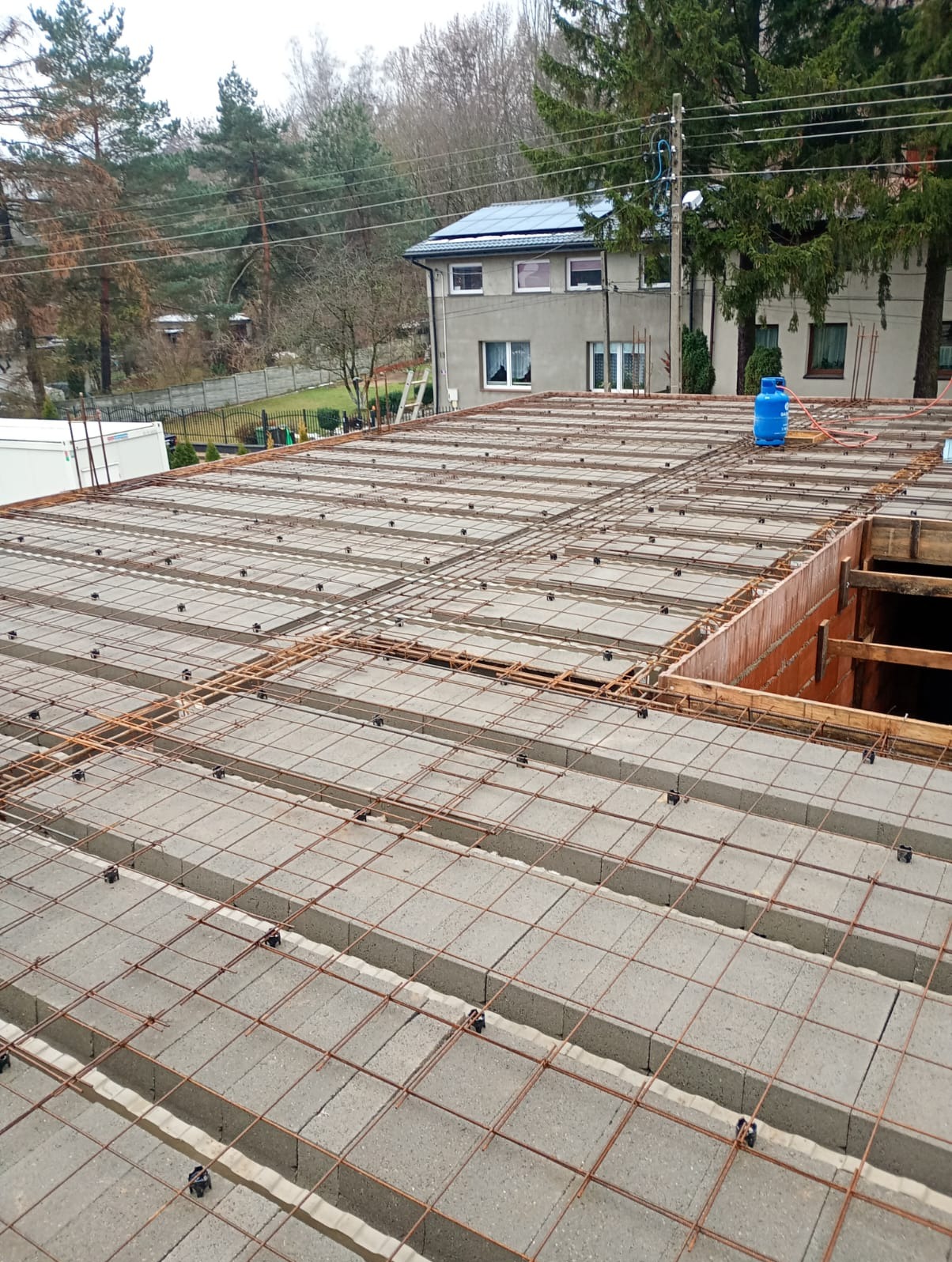 Zbrojony strop Teriva z pustaków szalunkowych, widok z góry na budowę domu jednorodzinnego. W tle dom z panelami słonecznymi i drzewa.