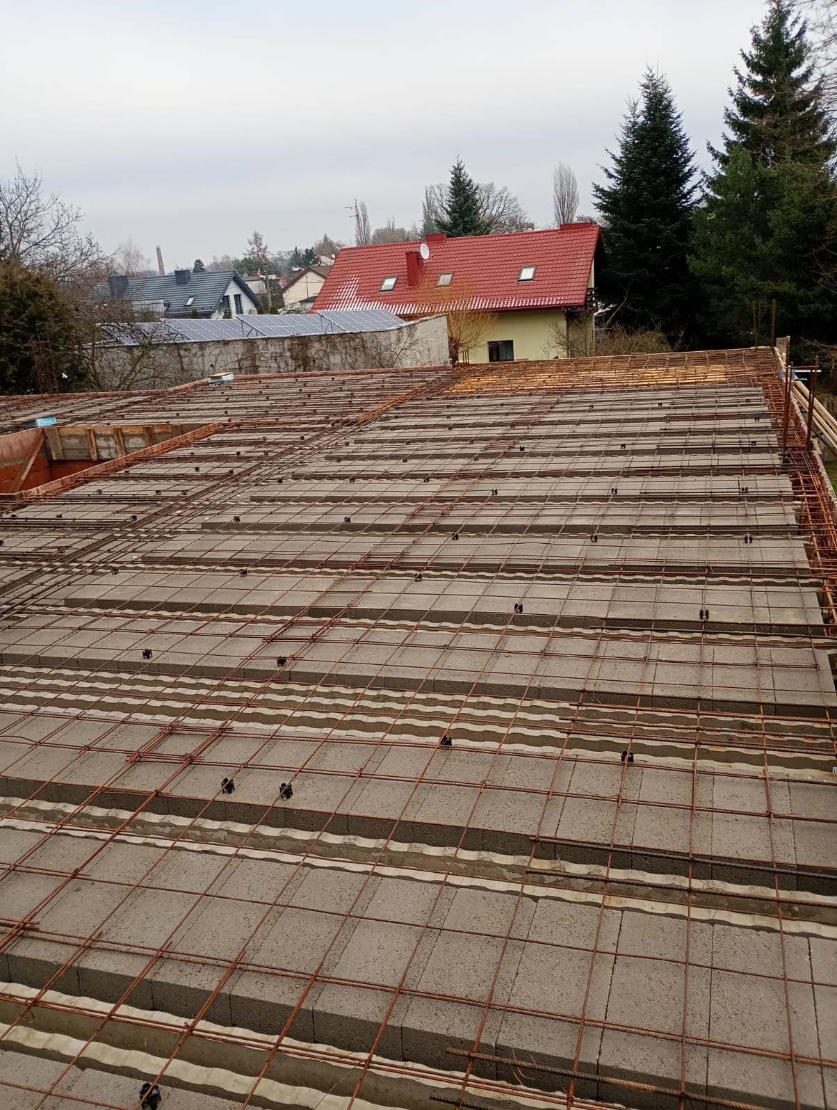 Zbrojenie stropu Teriva z betonowych pustaków szalunkowych, widoczne z góry na tle zabudowy jednorodzinnej w pochmurny dzień. Detal konstrukcji.