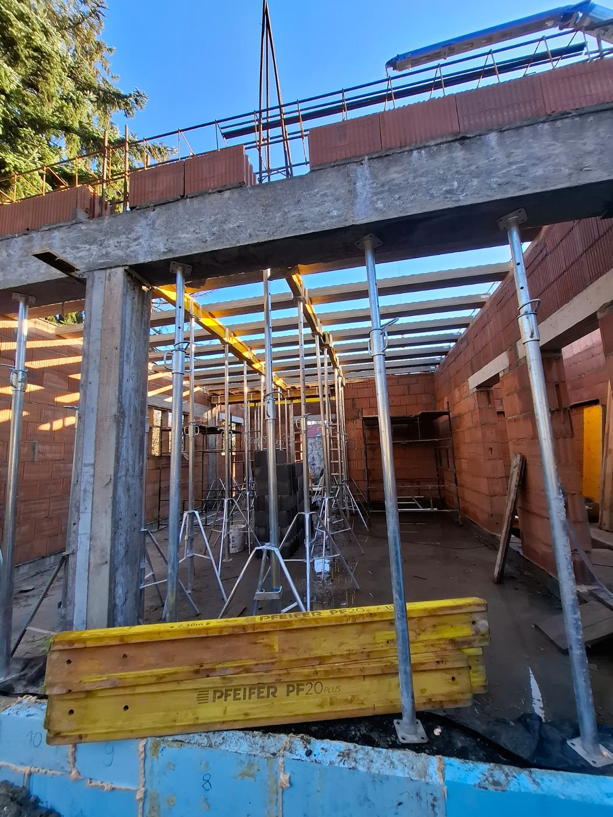 Wznoszenie ścian z czerwonej cegły na budowie domu. Widoczne podpory stropowe, szalunki i zbrojenia. Niebieskie niebo w tle.