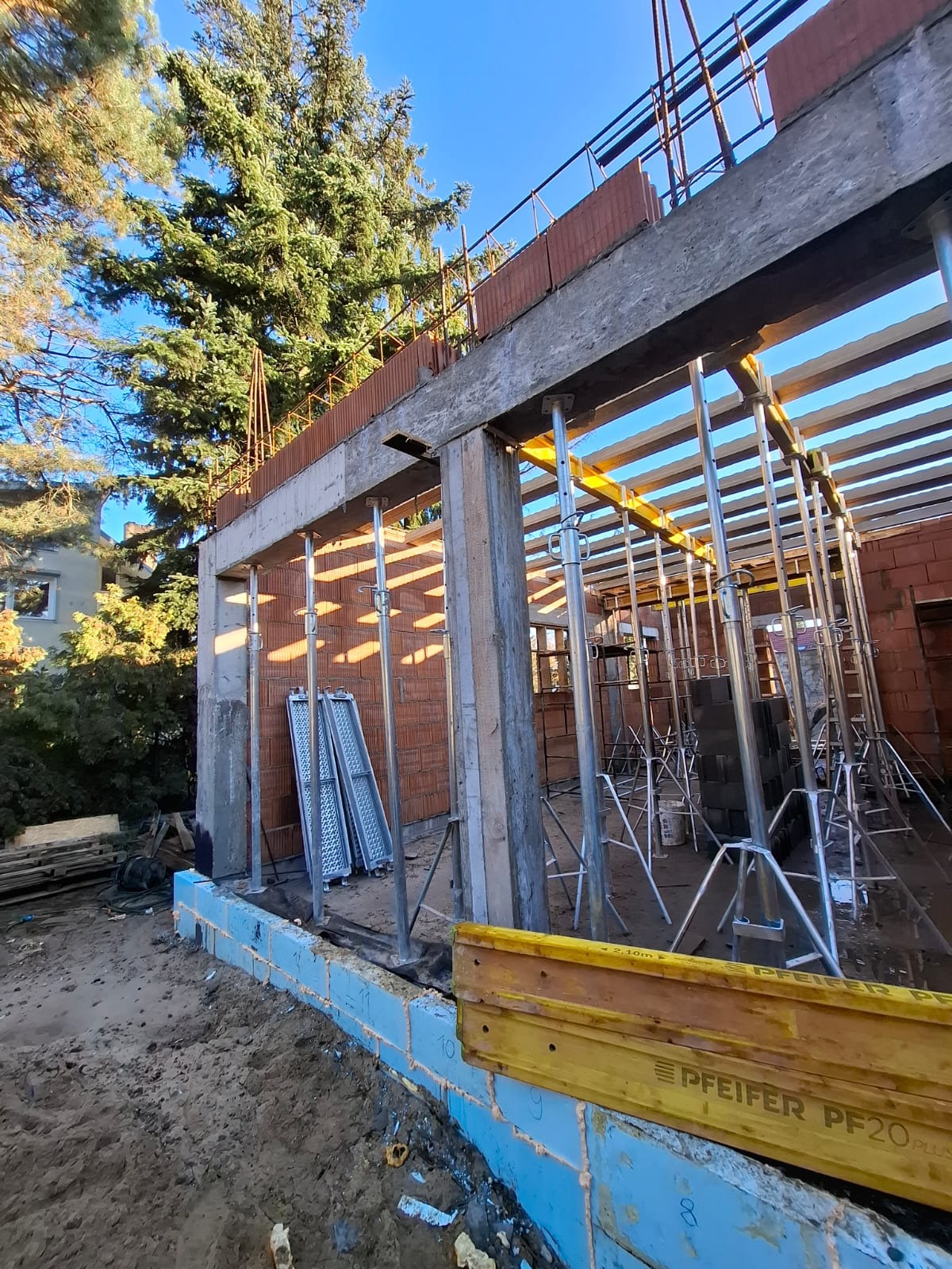 Surowy stan domu w trakcie budowy: ściany z cegły, betonowe słupy, podpory stalowe, szalunki. Widoczne elementy konstrukcyjne i materiały budowlane na tle drzew i błękitnego nieba.