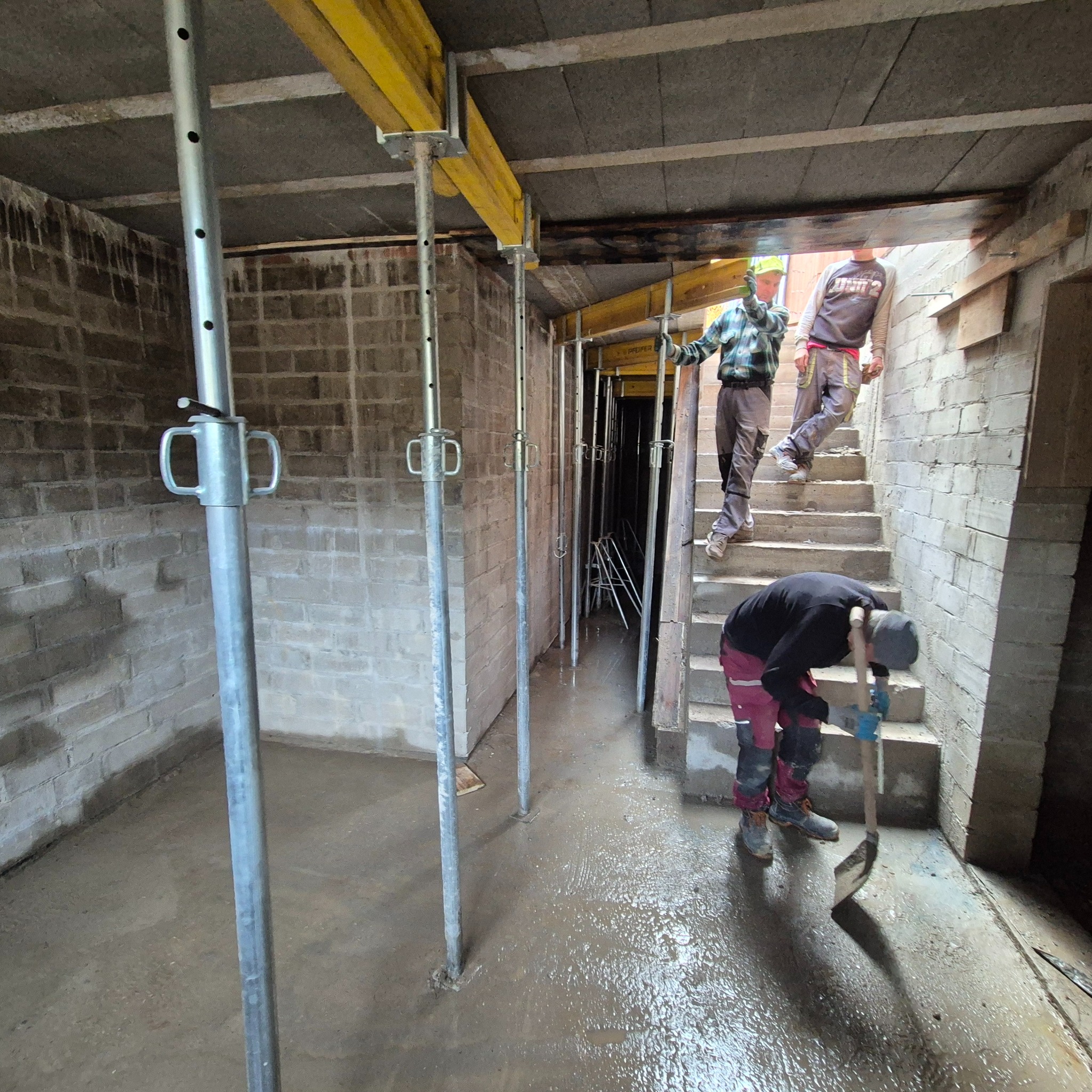 Wnętrze w trakcie wylewania posadzki. Widoczne podpory stropowe, pracownicy z łopatami i beton na podłodze. Surowe ściany z bloczków.