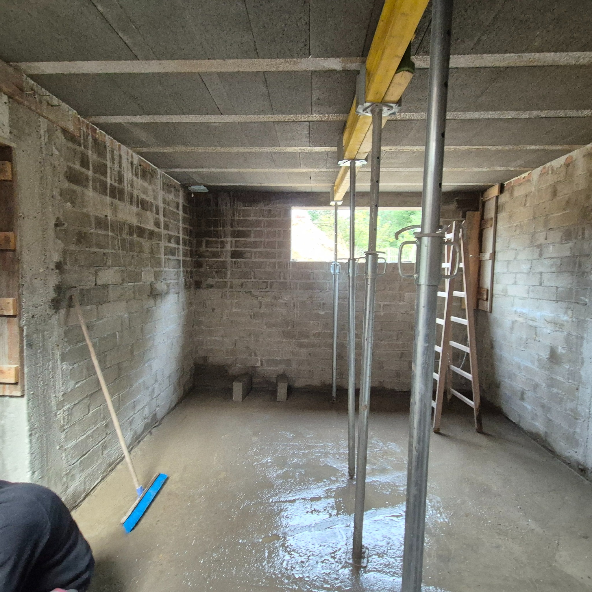 Surowe wnętrze z betonowymi ścianami i stropem, wsparte metalowymi stemplami. Widoczne okno, drabina i narzędzia. Podłoga pokryta świeżym betonem.