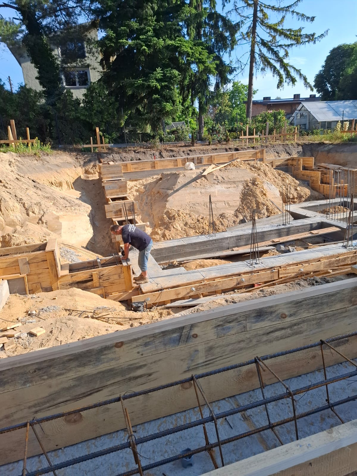 Budowa fundamentów domu: szalunki z desek, zbrojenie stalowe, wykop ziemny. Pracownik w tle. Widok z góry na plac budowy w słoneczny dzień.