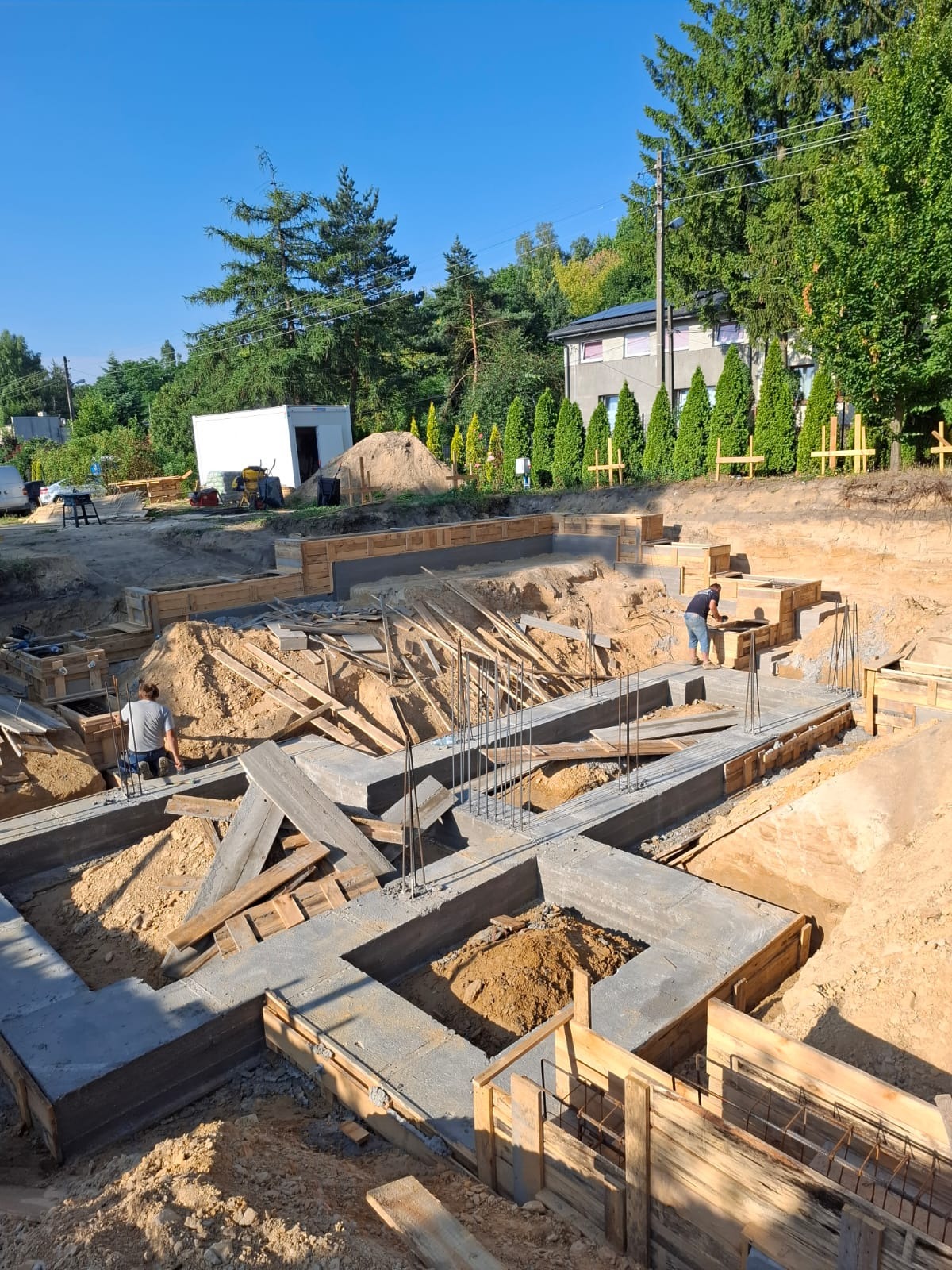Fundamenty domu w trakcie budowy: betonowe ściany szalunkowe, zbrojenia stalowe, drewniane elementy konstrukcyjne. Dwóch pracowników w trakcie prac. Widok z góry, słoneczny dzień.