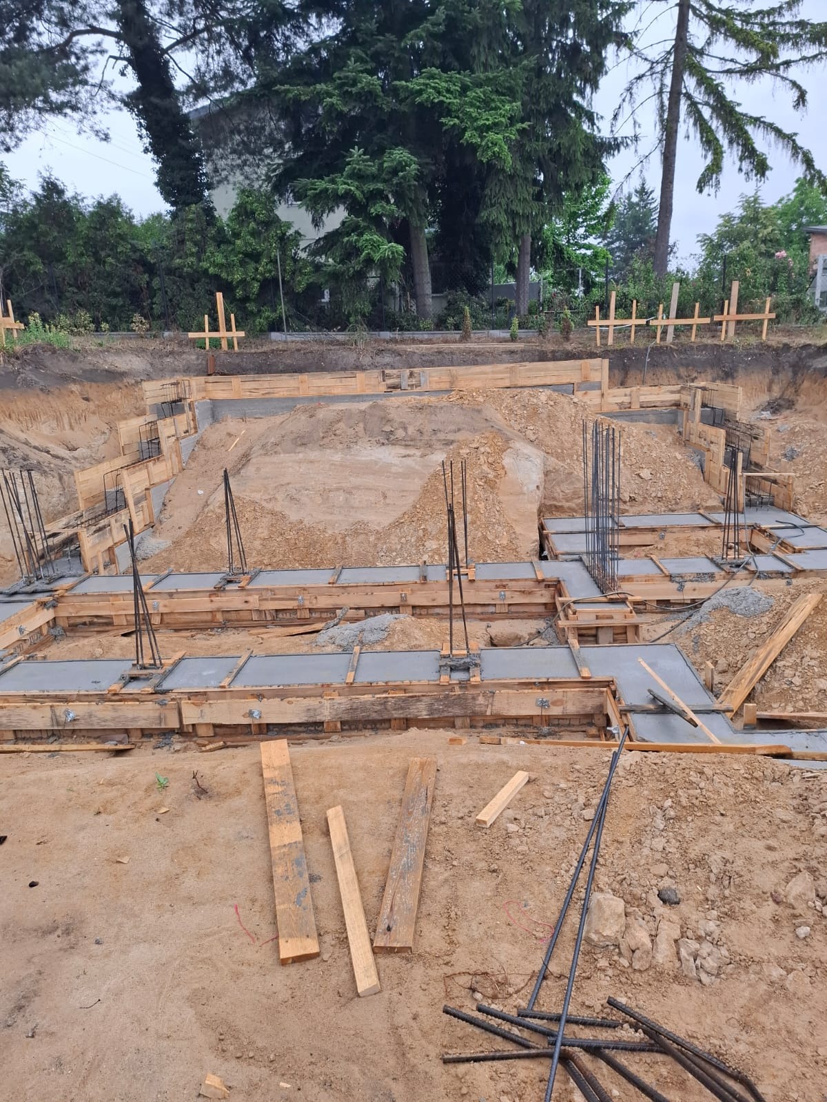 Wykop pod fundamenty domu z drewnianym szalunkiem i częściowo wylanym betonem. Widoczne wystające pręty zbrojeniowe. Tło: drzewa i częściowo widoczny budynek.