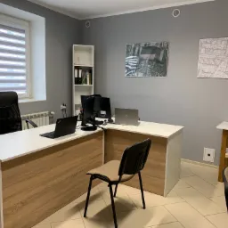 Biuro geodety z dwoma laptopami na białym biurku z drewnianym frontem, krzesłem, regałem i mapami na ścianie