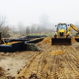 ArchiBud - Przygotowanie fundamentów pod budynek: wykopane rowy z izolacją, zbrojenia, pracownicy z zagęszczarką i koparka w tle, mglisty poranek.