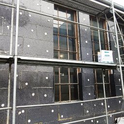 ArchiBud - Elewacja budynku w trakcie ocieplania czarnym styropianem, widoczne rusztowanie i pracownik w niebieskiej koszulce na górze, puszki z klejem na rusztowaniu, okna z brązowymi ramami.