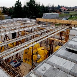 ArchiBud - Budowa stropu Teriva z pustaków szalunkowych, belek stalowych i żółtych paczek z materiałem izolacyjnym, widok z góry na plac budowy w trakcie dnia.