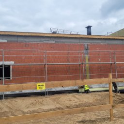 ArchiBud - Budynek w trakcie ocieplania, widoczne rusztowania, niezabezpieczony otwór okienny, fragment ocieplenia na ścianie, żółta tabliczka ostrzegawcza 'Uwaga! Głębokie wykopy'.