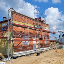 ArchiBud - Budynek w trakcie ocieplania elewacji z widocznymi rusztowaniami, izolacją z wełny mineralnej, paletami z materiałami izolacyjnymi i transformatorem w tle, w słoneczny dzień.
