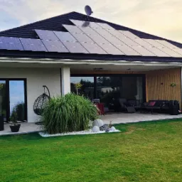 Nowoczesny dom jednorodzinny z panelami słonecznymi na dachu, widok z ogrodu na taras z meblami wypoczynkowymi i ozdobnymi trawami.