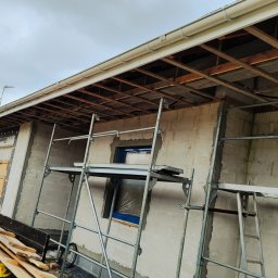 ArchiBud - Budynek w trakcie tynkowania elewacji, widoczne rusztowanie, odsłonięta konstrukcja dachu, oraz samochód dostawczy z deskami.