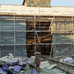 Budynek przemysłowy w trakcie ocieplania, widoczne rusztowania, płyty izolacyjne z kołkami, puszki z farbą i resztki materiałów budowlanych na ziemi, tory kolejowe w tle.