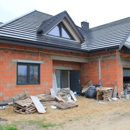 ArchiBud - Budynek w stanie surowym otwartym w Zduńskiej Woli, z widoczną konstrukcją z czerwonej cegły, czarną dachówką i oknami; na ziemi porozrzucane materiały budowlane i palety.