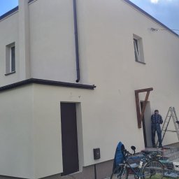 ArchiBud - Jasna elewacja domu jednorodzinnego po odświeżeniu. Dwóch pracowników na drabinie podczas prac wykończeniowych. Widoczny rower i wózek.