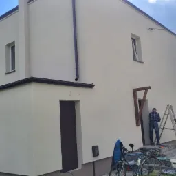 Jasna elewacja domu jednorodzinnego po odświeżeniu. Dwóch pracowników na drabinie podczas prac wykończeniowych. Widoczny rower i wózek.