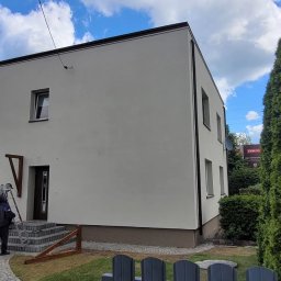 ArchiBud - Dwukondygnacyjny dom po tynkowaniu elewacji zewnętrznej, z pracownikiem przy schodach i narzędziami obok, w otoczeniu zieleni i błękitnego nieba.