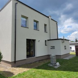 ArchiBud - Elewacja domu po tynkowaniu i malowaniu na jasny kolor. Widoczny fragment ogrodu, okna z czarnymi ramami i rury spustowe. Słoneczny dzień.
