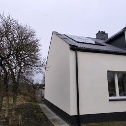 ArchiBud - Ściana domu po tynkowaniu z widoczną elewacją w kolorze białym, czarną podmurówką i panelami słonecznymi na dachu. Obok drzewo bez liści i fragment ogrodu.