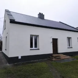 Biały dom jednorodzinny z szarą blachodachówką i ciemnym cokołem. Widoczne okna z białymi ramami i brązowe drzwi wejściowe. Dom po remoncie elewacji.