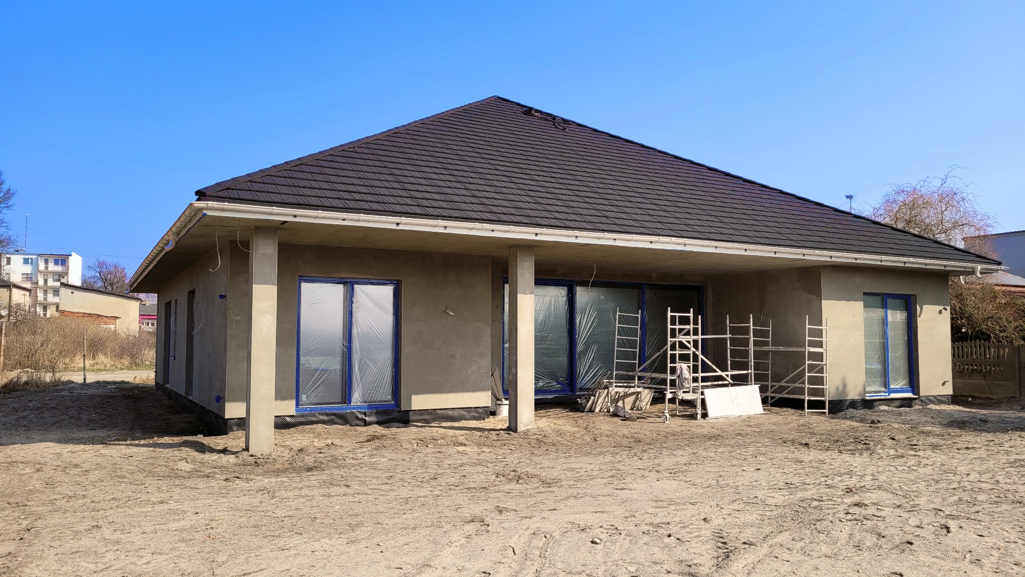 Dom jednorodzinny w trakcie prac elewacyjnych, okna zabezpieczone folią, widoczne rusztowania, świeżo położony tynk zewnętrzny, czarny dach z blachodachówki.