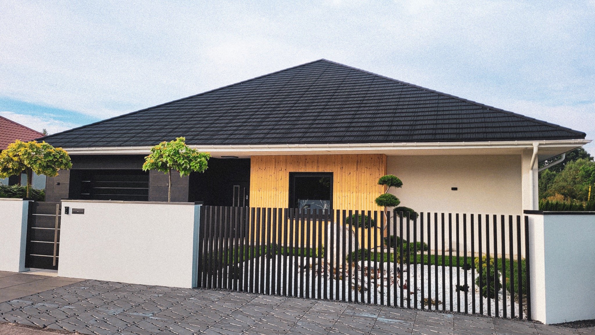 Nowoczesny dom jednorodzinny z czarnym dachem skośnym, jasną elewacją, drewnianą okładziną i minimalistycznym ogrodzeniem z pionowych sztachet.