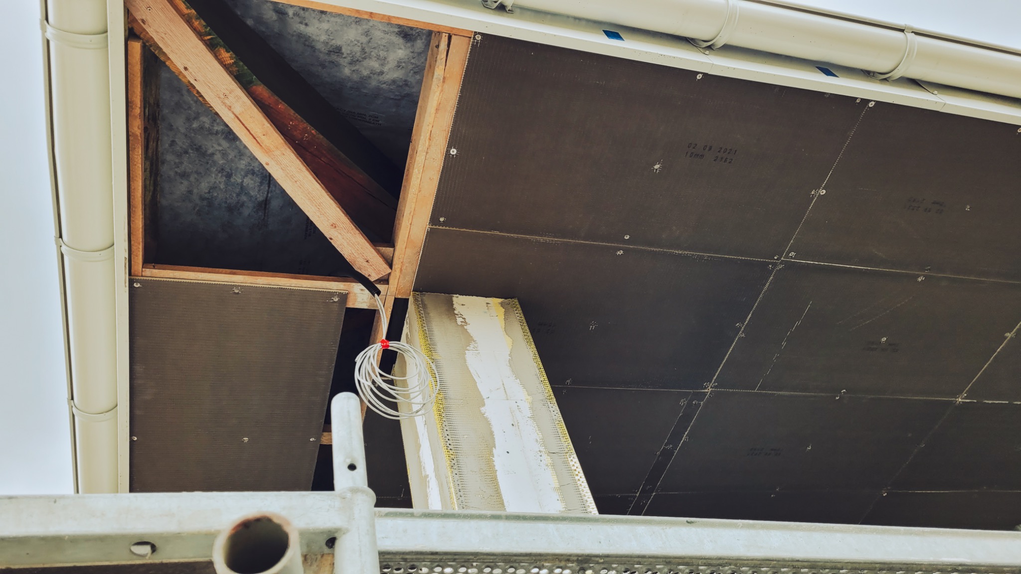 Montaż paneli izolacyjnych na podbitce dachu, widoczne drewniane elementy konstrukcyjne i fragment rusztowania.