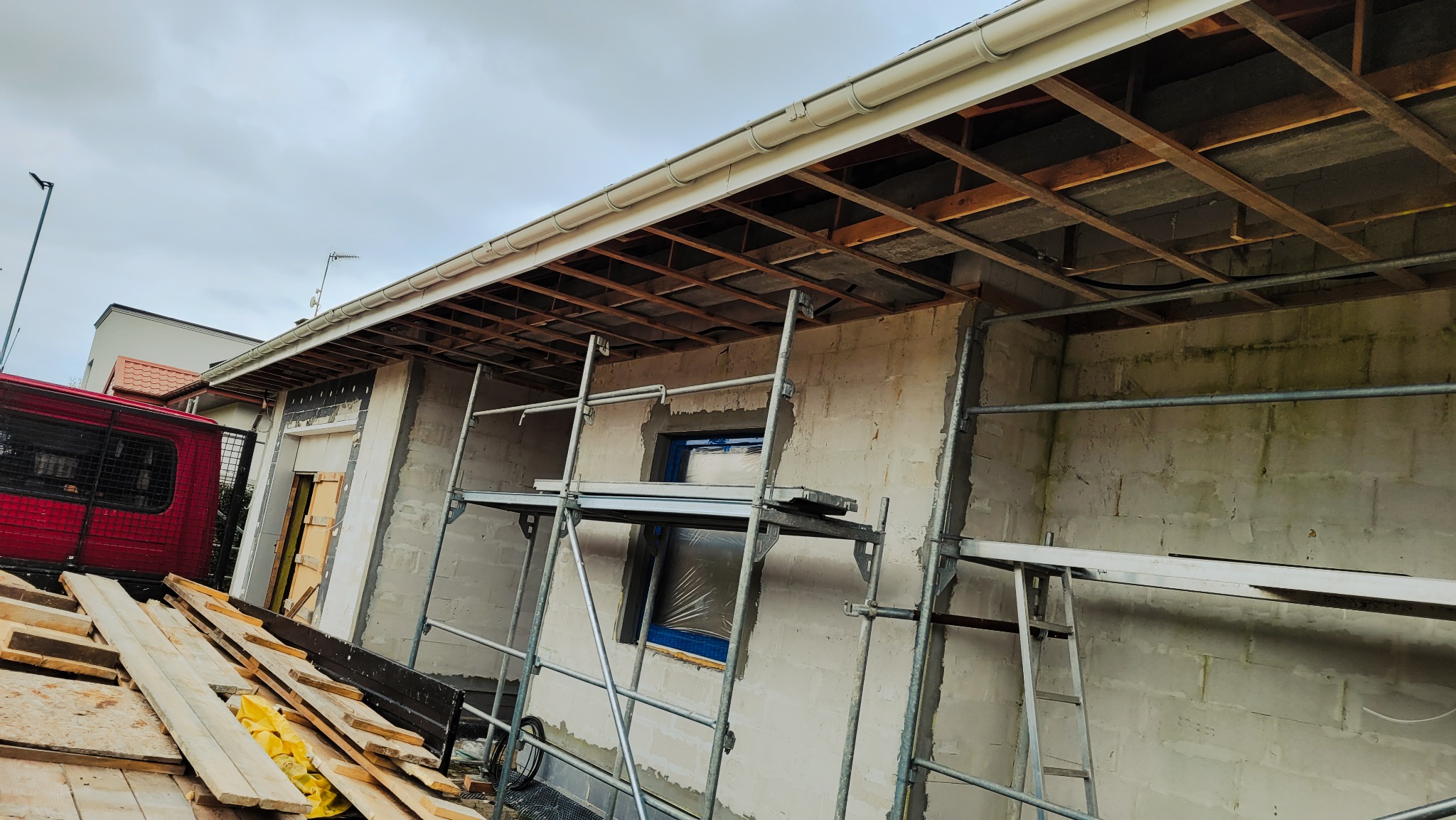 Budynek w trakcie tynkowania elewacji, widoczne rusztowanie, odsłonięta konstrukcja dachu, oraz samochód dostawczy z deskami.