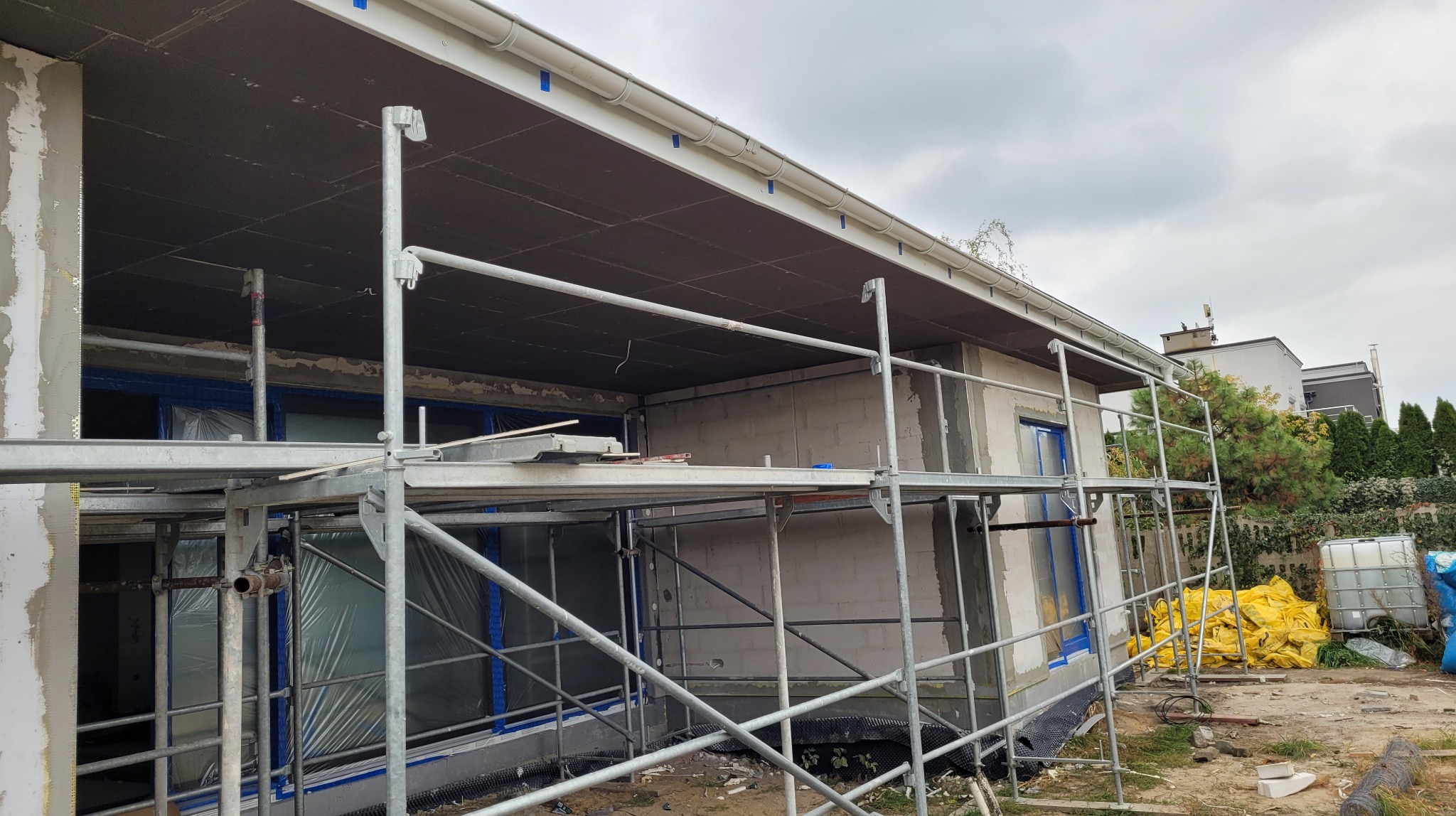 Dom jednorodzinny w trakcie ocieplania elewacji, widoczne rusztowanie, zabezpieczone okna folią, ocieplony fragment podbitki dachu