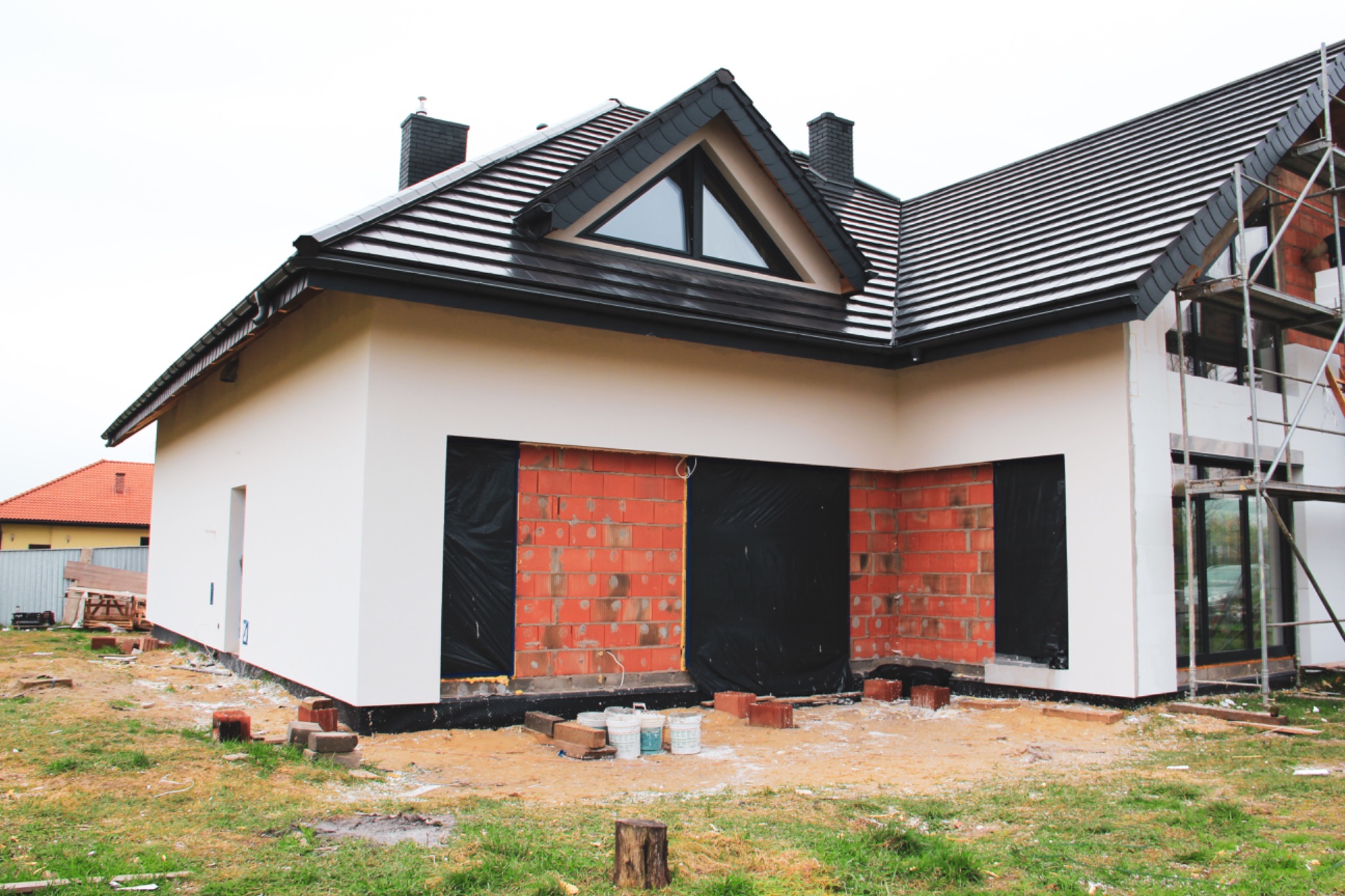 Dom jednorodzinny w trakcie prac wykończeniowych elewacji: częściowo otynkowana i pomalowana ściana, fragmenty z cegły z czarną folią zabezpieczającą, rusztowanie przy oknie, kilka wiader z farbą...