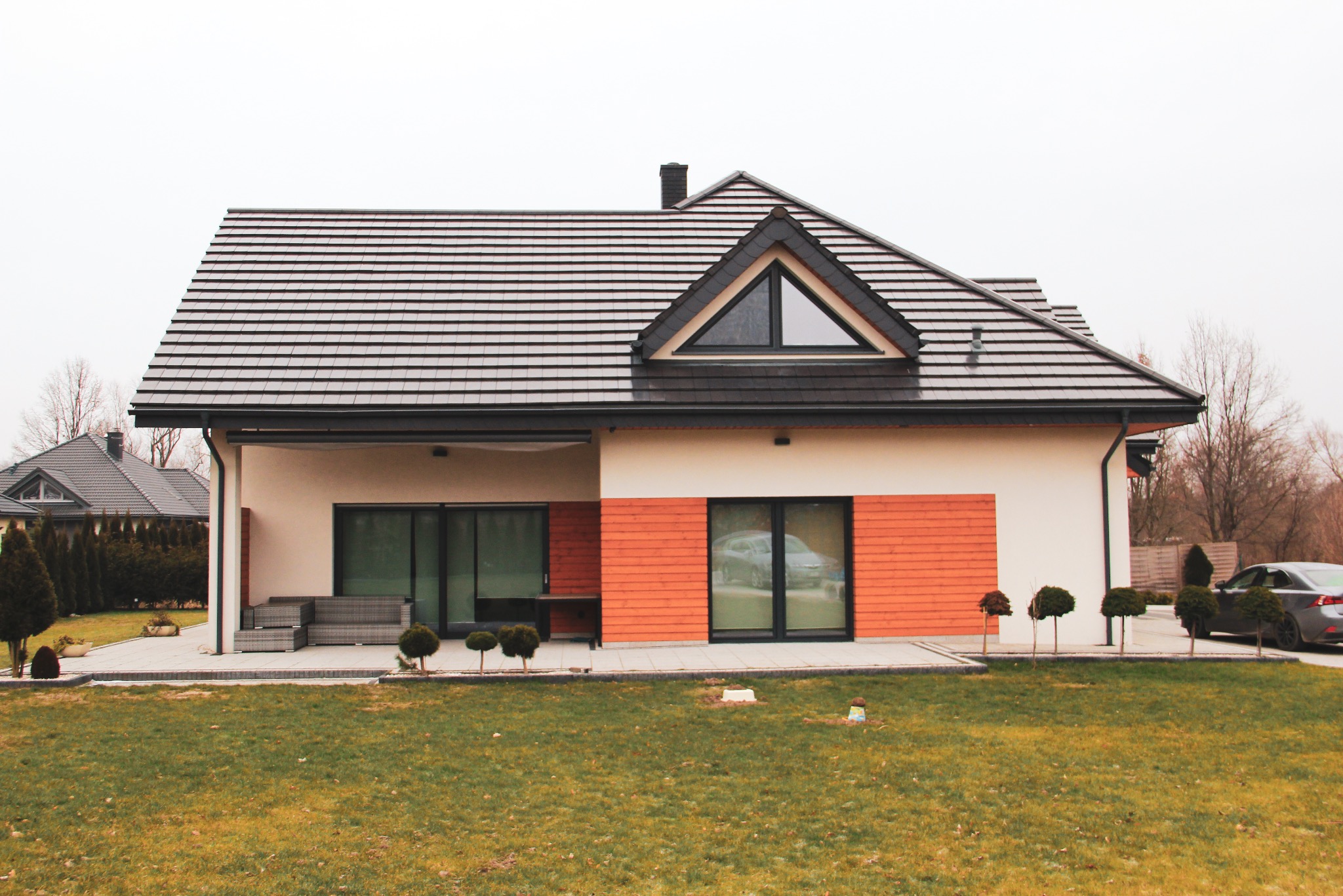 Nowoczesny dom jednorodzinny z dwuspadowym dachem pokrytym ciemną dachówką, jasną elewacją z elementami drewnianej okładziny w kolorze ceglastym, duże przesuwne okna i zadbany trawnik...