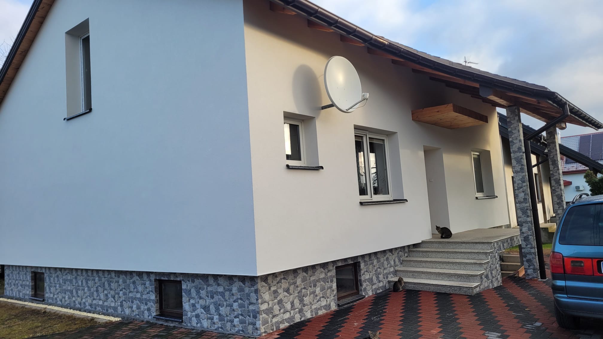 Biała elewacja domu jednorodzinnego z szarą podmurówką i czarnym kotem siedzącym na schodach wejściowych. Widoczna antena satelitarna i fragment niebieskiego samochodu.