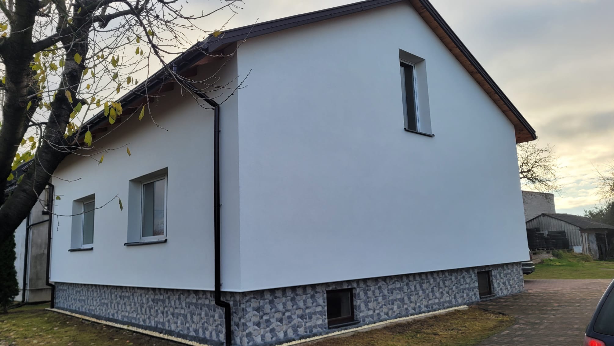 Biały dom z szarą elewacją przy gruncie i brązowym dachem. Widoczne dwa okna na parterze i jedno na poddaszu. W tle drzewo i budynki gospodarcze.