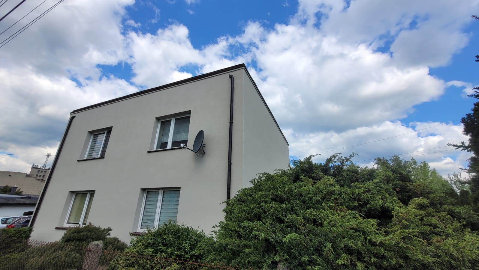 Dwupiętrowy dom z jasną elewacją, ciemnymi detalami i anteną satelitarną na tle błękitnego nieba z chmurami. Widok z dołu, zieleń w kadrze.