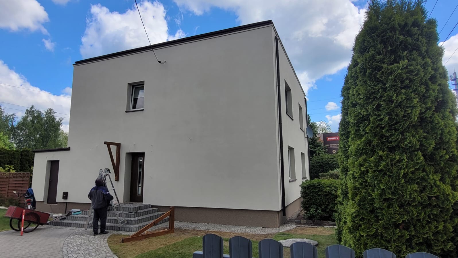 Dwukondygnacyjny dom po tynkowaniu elewacji zewnętrznej, z pracownikiem przy schodach i narzędziami obok, w otoczeniu zieleni i błękitnego nieba.